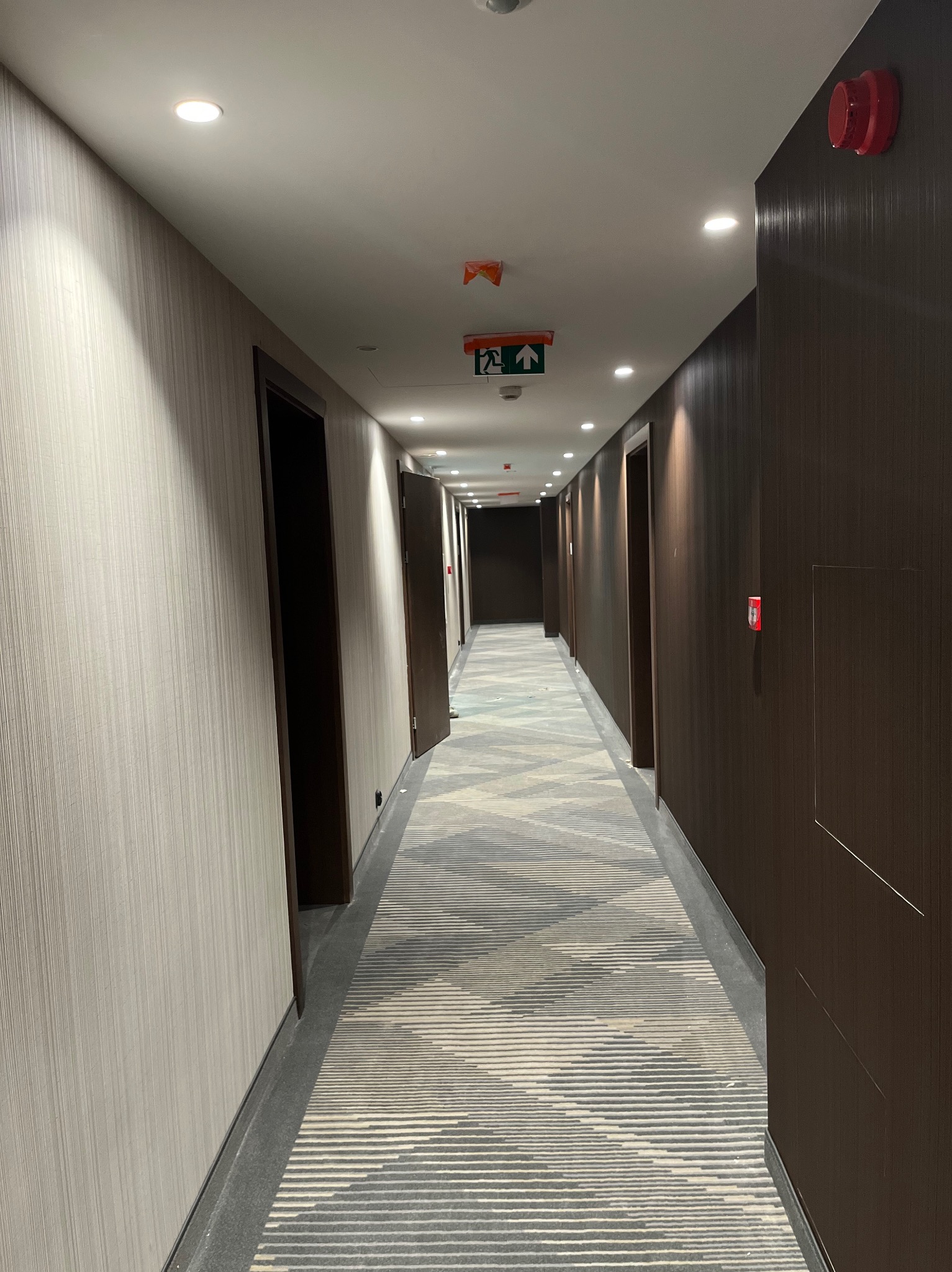 Długi korytarz hotelowy z tapetowanymi ścianami w odcieniach szarości i brązu, wzorzysta wykładzina podłogowa, oświetlenie sufitowe i drzwi po obu stronach.