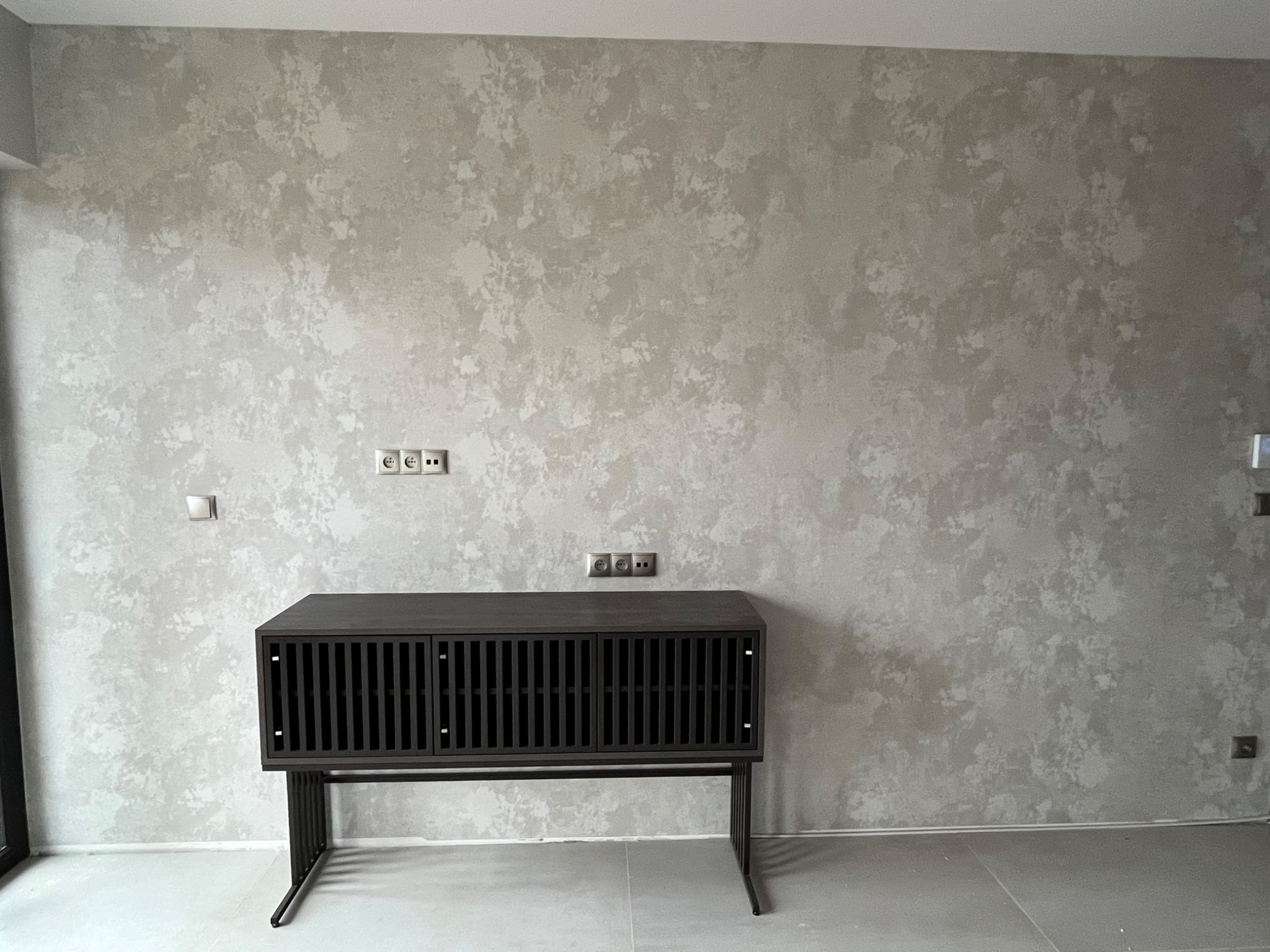 Ściana z dekoracyjną, szarą fakturą przypominającą chmurki, pod nią ciemna szafka RTV z lamelami.