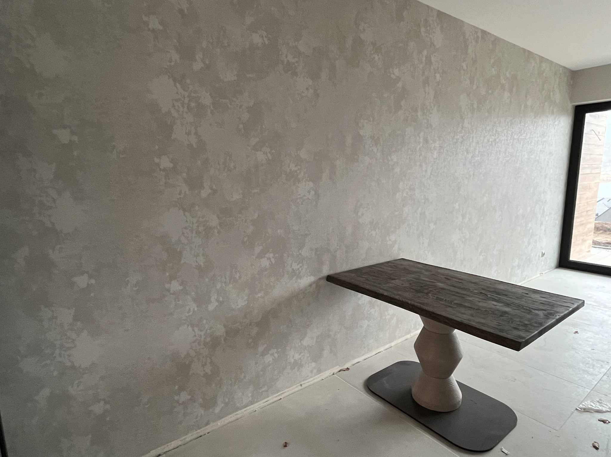 Ściana z dekoracyjną, beżową fakturą przypominającą marmur, widoczny fragment nowoczesnego stołu na tle okna.