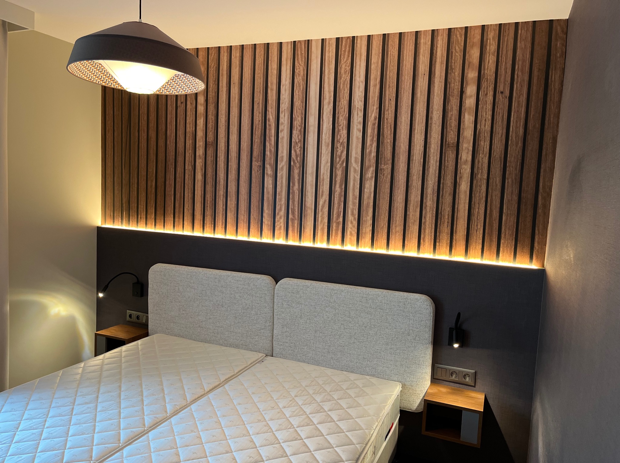 Nowoczesna sypialnia z drewnianą ścianą z lameli podświetloną od tyłu, szara tapicerowana rama łóżka i minimalistyczne lampki nocne.