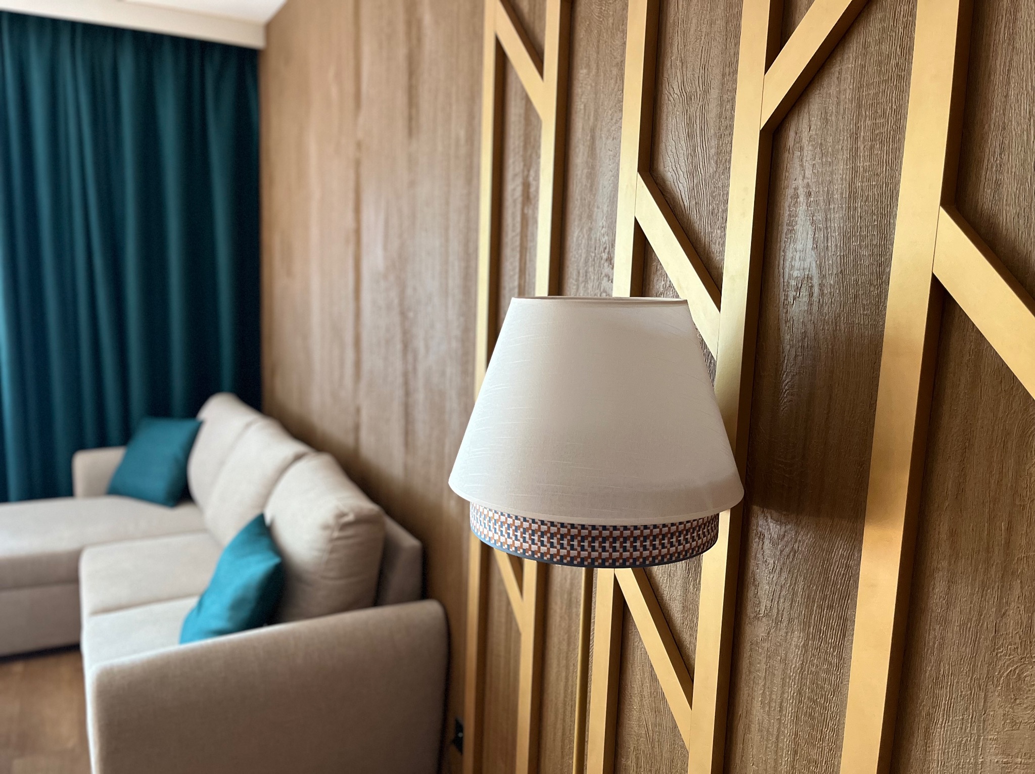 Nowoczesny salon z drewnianą ścianą dekoracyjną w geometryczne złote ramy, lampą ścienną z wzorzystym abażurem i beżową sofą z turkusowymi poduszkami.