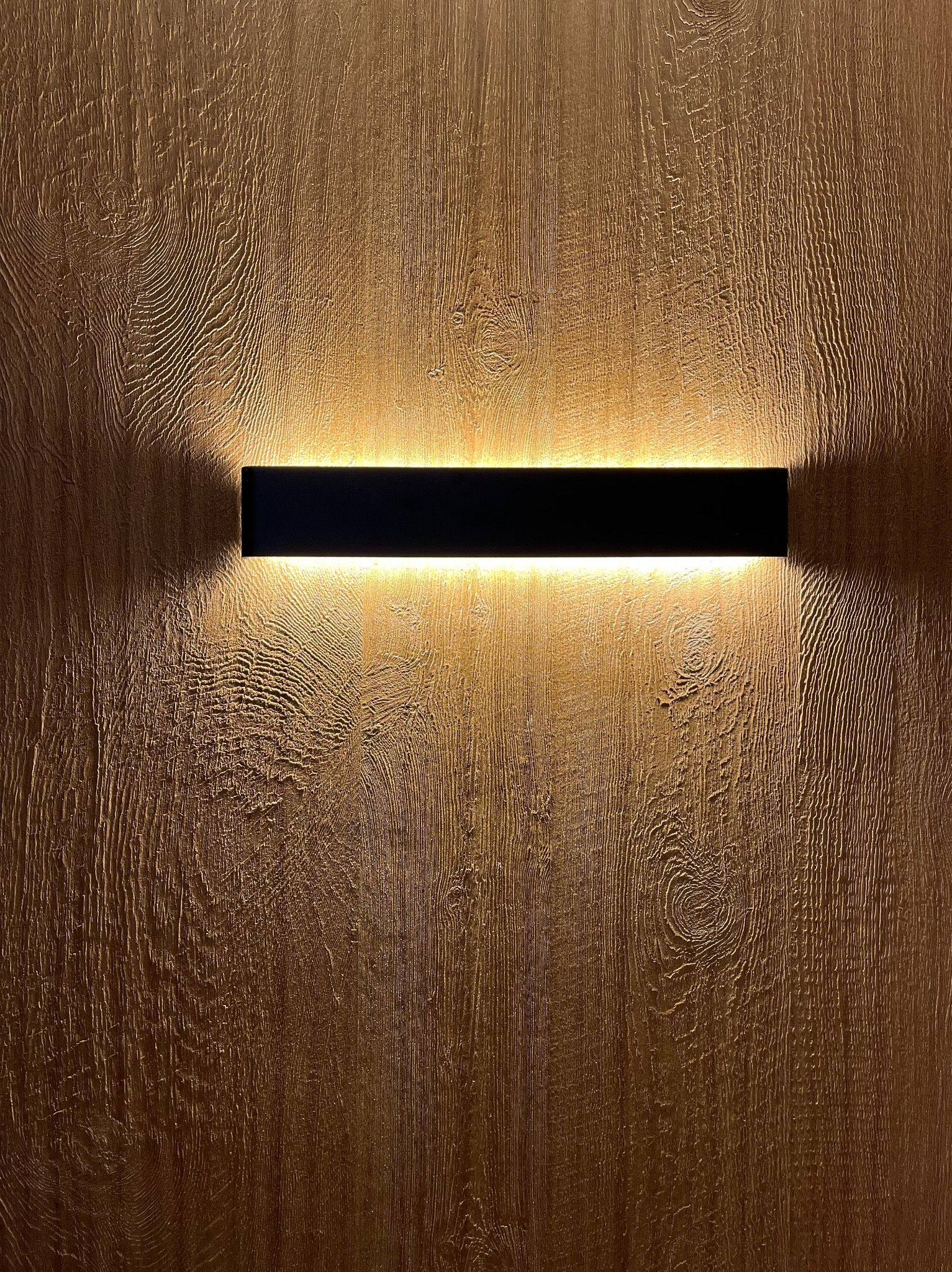 Dekoracyjna lampa ścienna LED o prostokątnym kształcie, zamontowana na ścianie z fakturą drewna, oświetlająca ją ciepłym światłem.