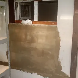 Ściana z częściowo nałożoną warstwą szpachli w pomieszczeniu z oknem, widoczne przygotowania do dalszych prac wykończeniowych.