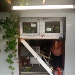 Montaż dwukomorowego okna PCV z naklejkami producenta, widoczny fragment ściany z bluszczem, narzędzia i materiały budowlane w tle, pracownik w rękawicach ochronnych