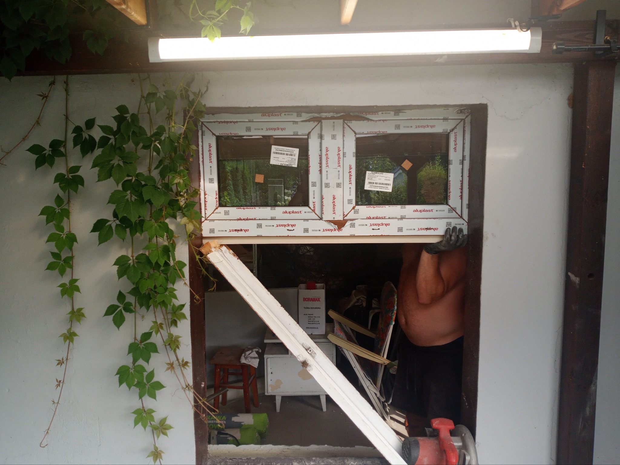 Montaż dwukomorowego okna PCV z naklejkami producenta, widoczny fragment ściany z bluszczem, narzędzia i materiały budowlane w tle, pracownik w rękawicach ochronnych