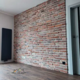 Dekoracyjna ściana z cegły klinkierowej w salonie, z widocznym grzejnikiem panelowym i fragmentem drzwi z szarą ościeżnicą, podłoga z paneli imitujących ciemne drewno.