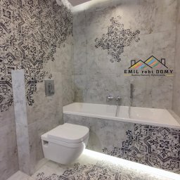 Emil Kliza - Emil robi Domy - Wykończona łazienka z białą armaturą, dekoracyjnymi płytkami ściennymi i podłogowymi w odcieniach szarości z czarnymi wzorami, podświetlona listwą LED pod wanną, widoczny podtynkowy system...