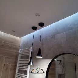 Emil Kliza - Emil robi Domy - Nowoczesna łazienka z drewnopodobnymi płytkami ułożonymi w jodełkę na podłodze i ścianie, oświetlona dwoma czarnymi lampami wiszącymi i ukrytym oświetleniem LED w suficie, z grzejnikiem drabinkowym...