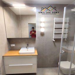 Emil Kliza - Emil robi Domy - Wykończona łazienka w szarościach z drewnianym blatem pod umywalką, kabiną prysznicową z widoczną toaletą oraz grzejnikiem drabinkowym; w lustrze odbija się osoba robiąca zdjęcie.