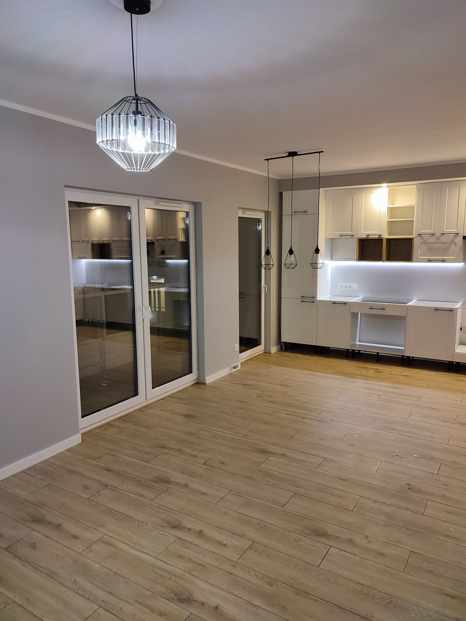 Nowoczesna kuchnia z białymi szafkami, drewnopodobną podłogą i designerskim oświetleniem, widok na przeszklone drzwi balkonowe.