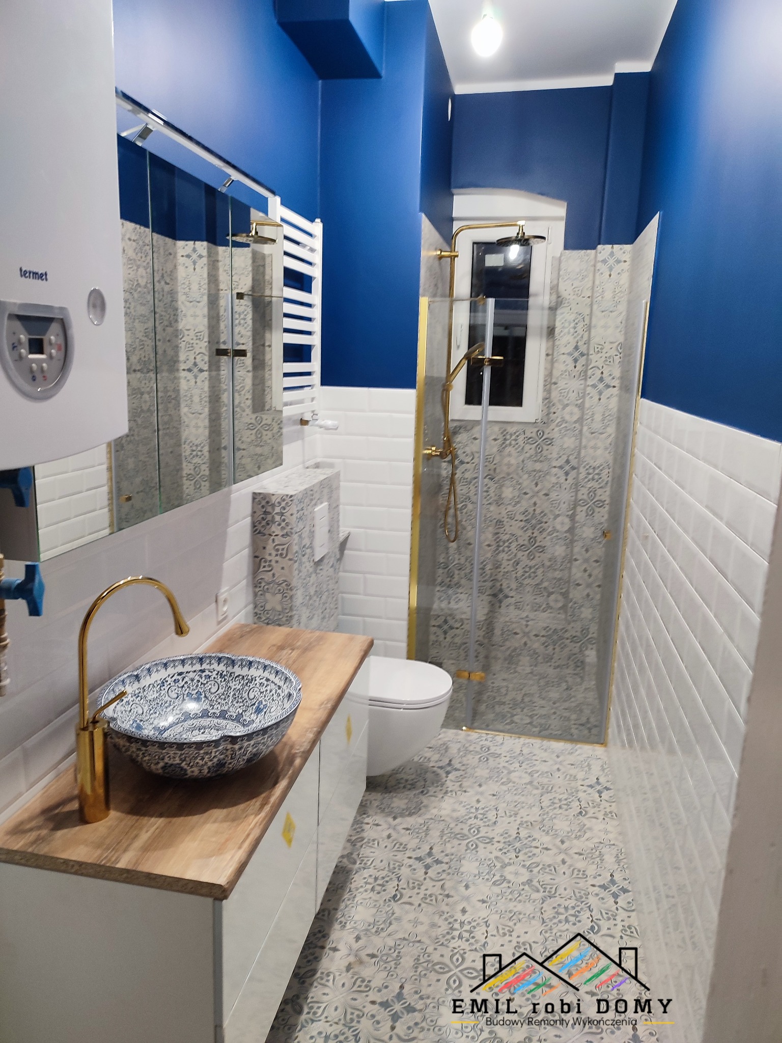 Niebiesko-biała łazienka z kabiną prysznicową w złotej ramie, umywalką w stylu orientalnym na drewnianym blacie, białą armaturą i grzejnikiem drabinkowym, oświetlona żarówką.