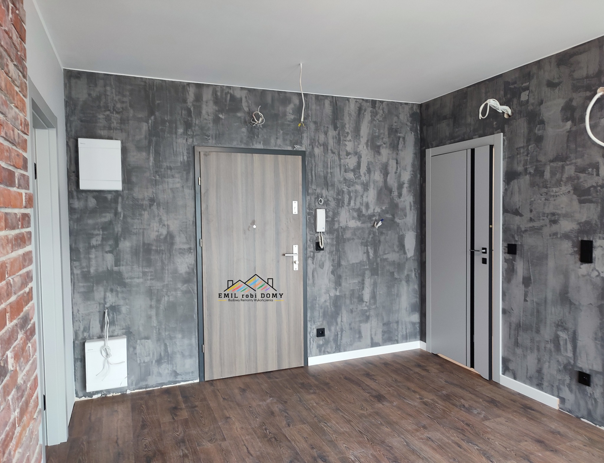 Wnętrze po remoncie: ściana z efektem betonu, dwoje drzwi (zewnętrzne i wewnętrzne), brązowe panele podłogowe, widoczne kable zwisające z sufitu, logo firmy na drzwiach wejściowych.