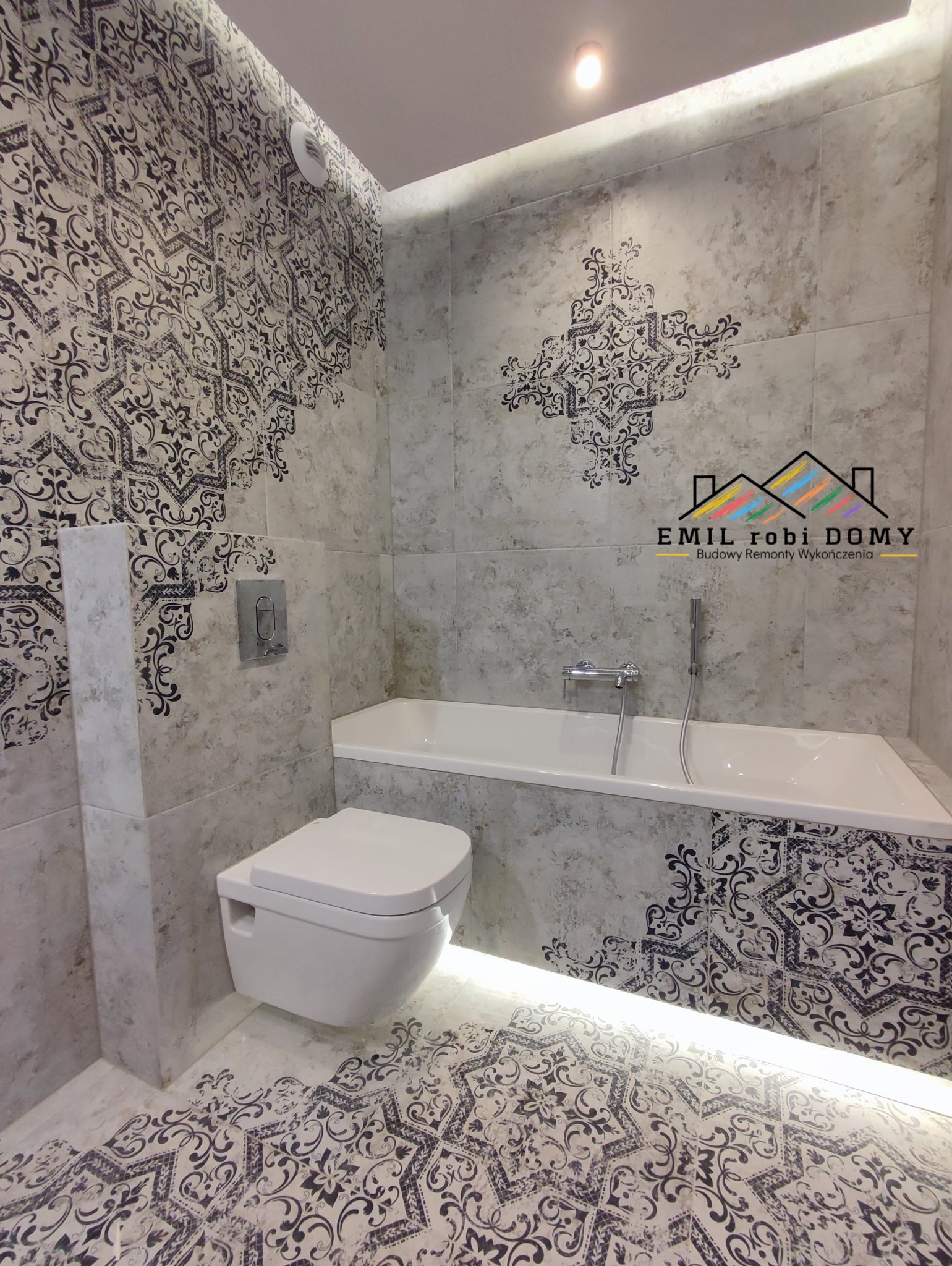 Wykończona łazienka z białą armaturą, dekoracyjnymi płytkami ściennymi i podłogowymi w odcieniach szarości z czarnymi wzorami, podświetlona listwą LED pod wanną, widoczny podtynkowy system...