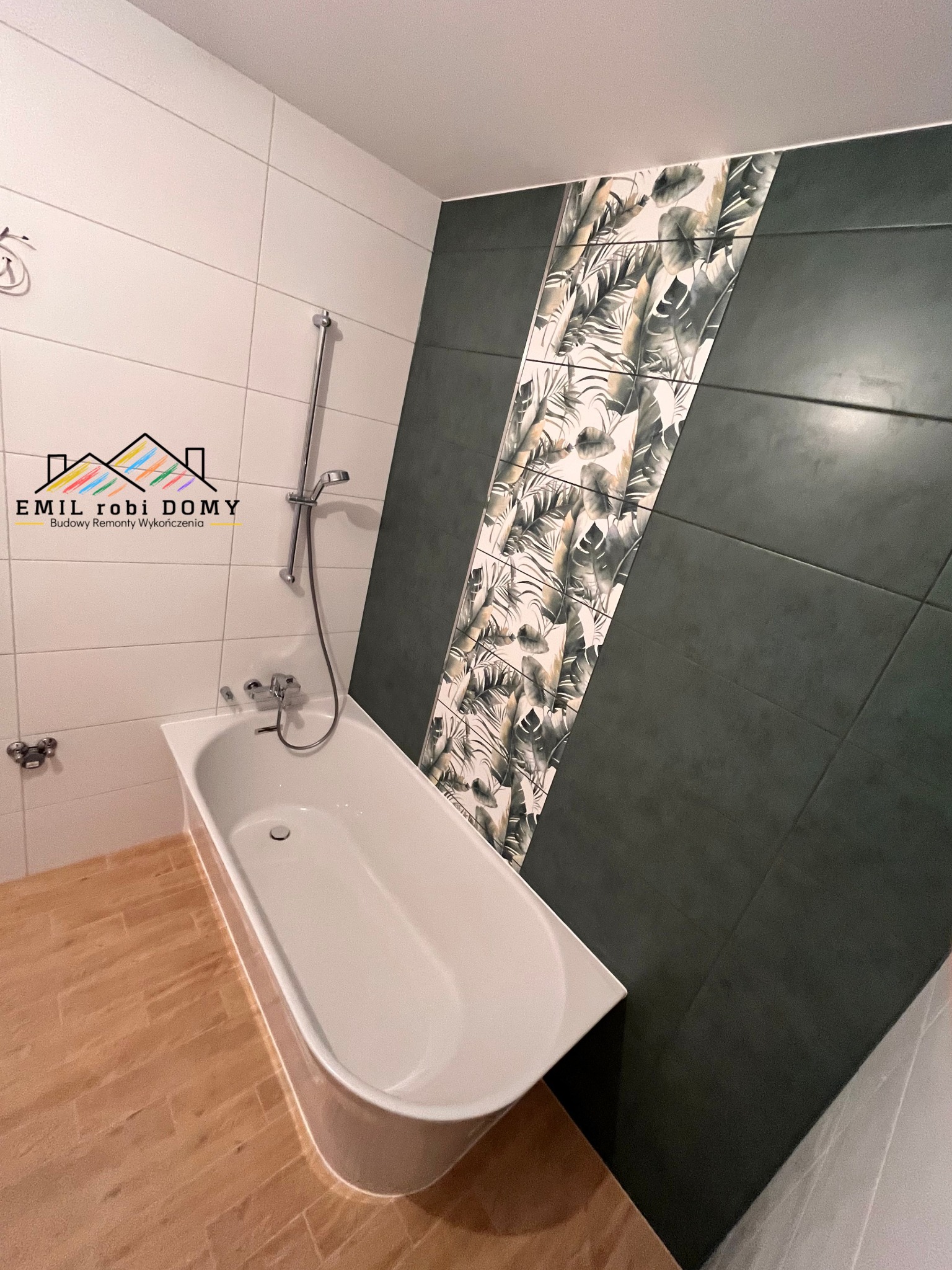 Biała wanna z chromowaną armaturą na tle wykafelkowanej ściany z motywem roślinnym, brązowa podłoga imitująca drewno, widoczne logo firmy budowlanej.