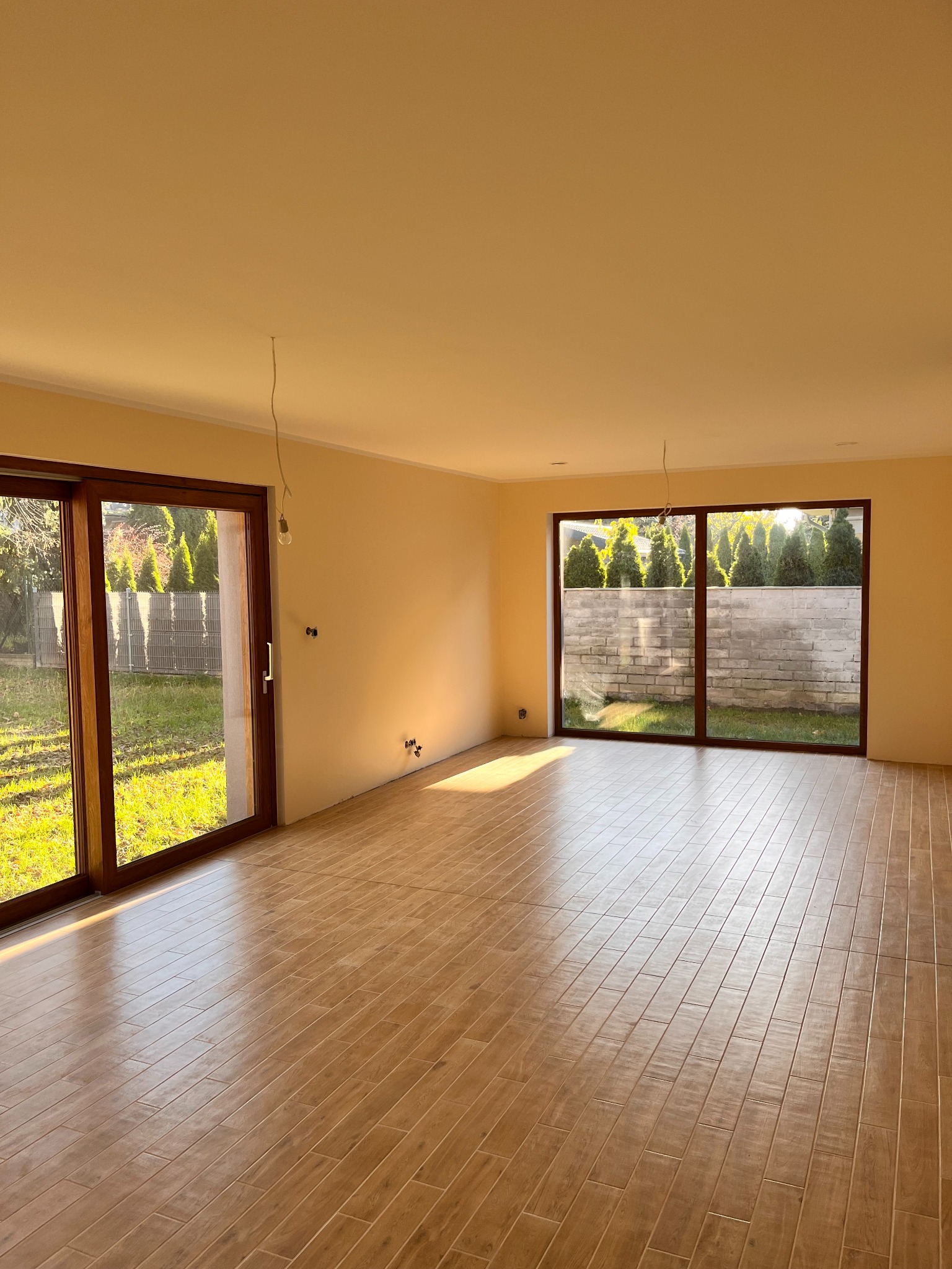 Jasne wnętrze z drewnopodobną podłogą, dużymi przesuwnymi drzwiami balkonowymi i oknem z widokiem na ogród, świeżo pomalowane ściany, widoczne wyprowadzenia elektryczne.