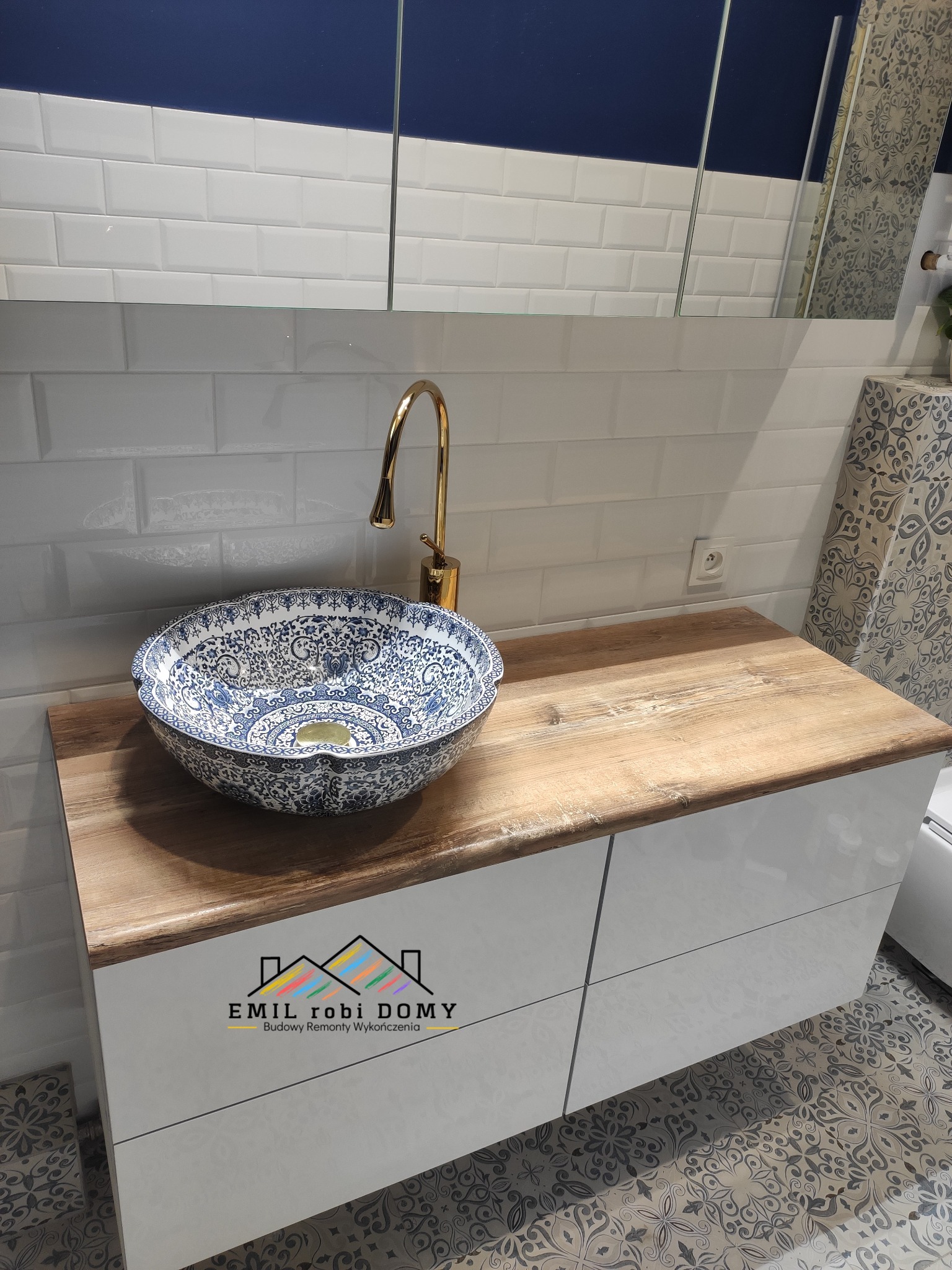 Umywalka nablatowa w stylu vintage, zdobiona niebieskim wzorem, zamontowana na drewnianym blacie szafki łazienkowej z białymi frontami, złota bateria, białe płytki na ścianie i wzorzysta podłoga.