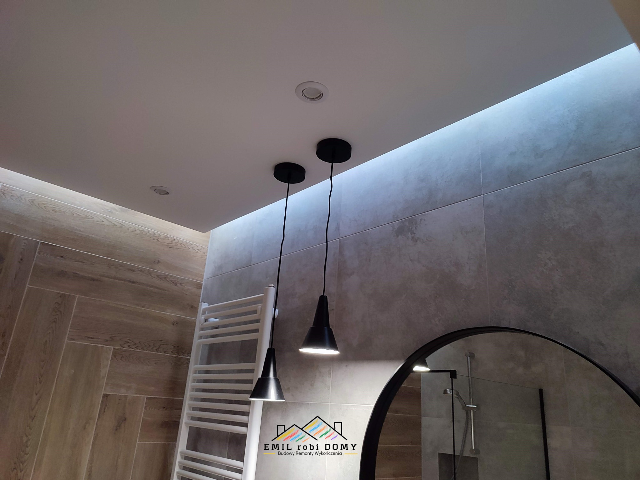 Nowoczesna łazienka z drewnopodobnymi płytkami ułożonymi w jodełkę na podłodze i ścianie, oświetlona dwoma czarnymi lampami wiszącymi i ukrytym oświetleniem LED w suficie, z grzejnikiem drabinkowym...