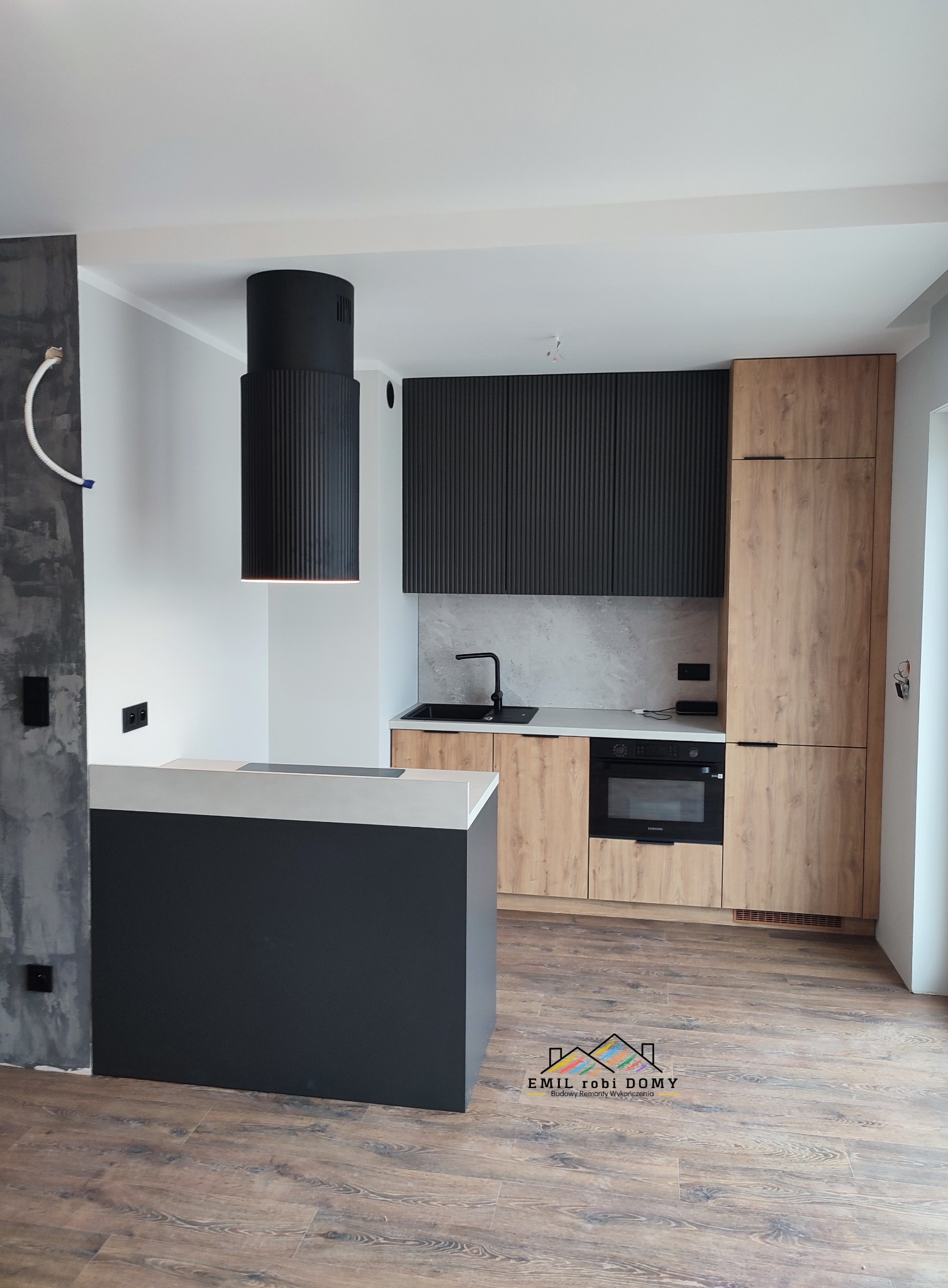 Nowoczesna kuchnia z wyspą, czarnym okapem cylindrycznym i drewnopodobnymi frontami szafek, widoczny fragment surowej ściany z instalacją elektryczną.