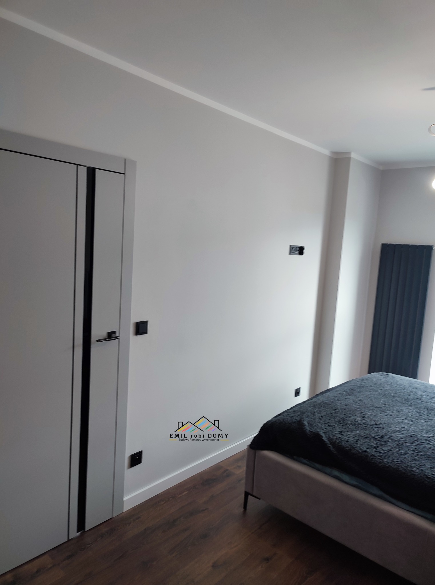 Nowoczesne wnętrze sypialni z szarą ścianą, ciemną podłogą, białymi listwami przypodłogowymi, szarymi drzwiami z czarnymi wstawkami i logo firmy budowlanej w rogu.