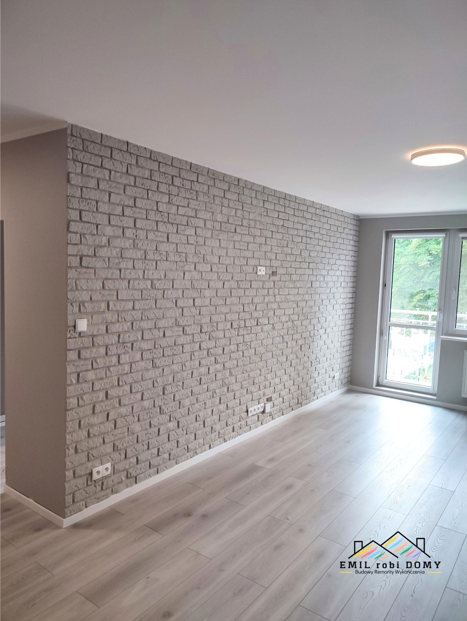 Ściana wykończona dekoracyjną cegłą w szarym odcieniu, z widocznymi gniazdkami elektrycznymi, na tle jasnej podłogi z paneli i okna z widokiem na zieleń.