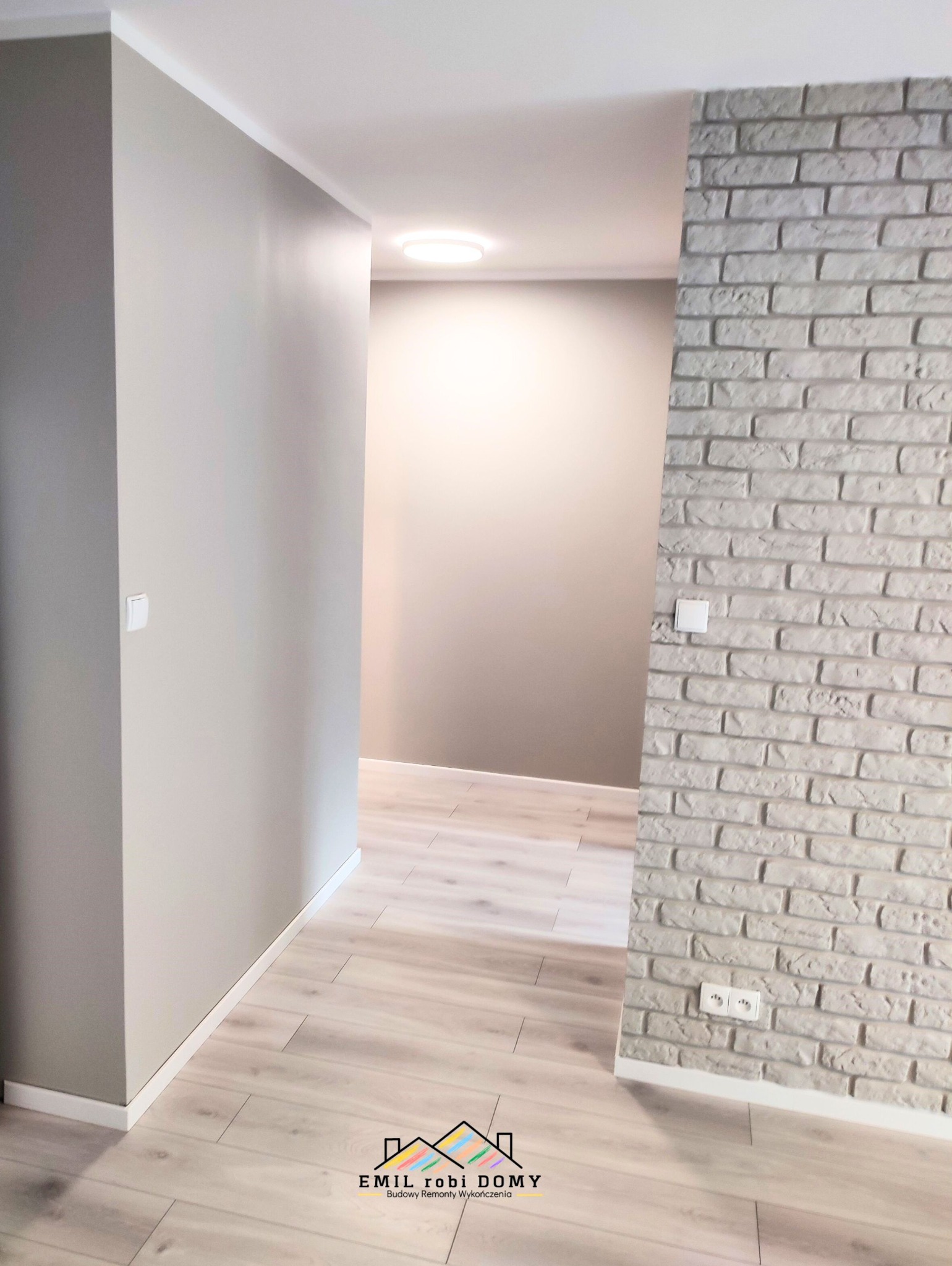 Korytarz po remoncie z szarą ścianą z cegły, gładkimi ścianami w kolorze beżowym i podłogą z paneli w odcieniach bieli i szarości; widoczny włącznik światła i podwójne gniazdko elektryczne.