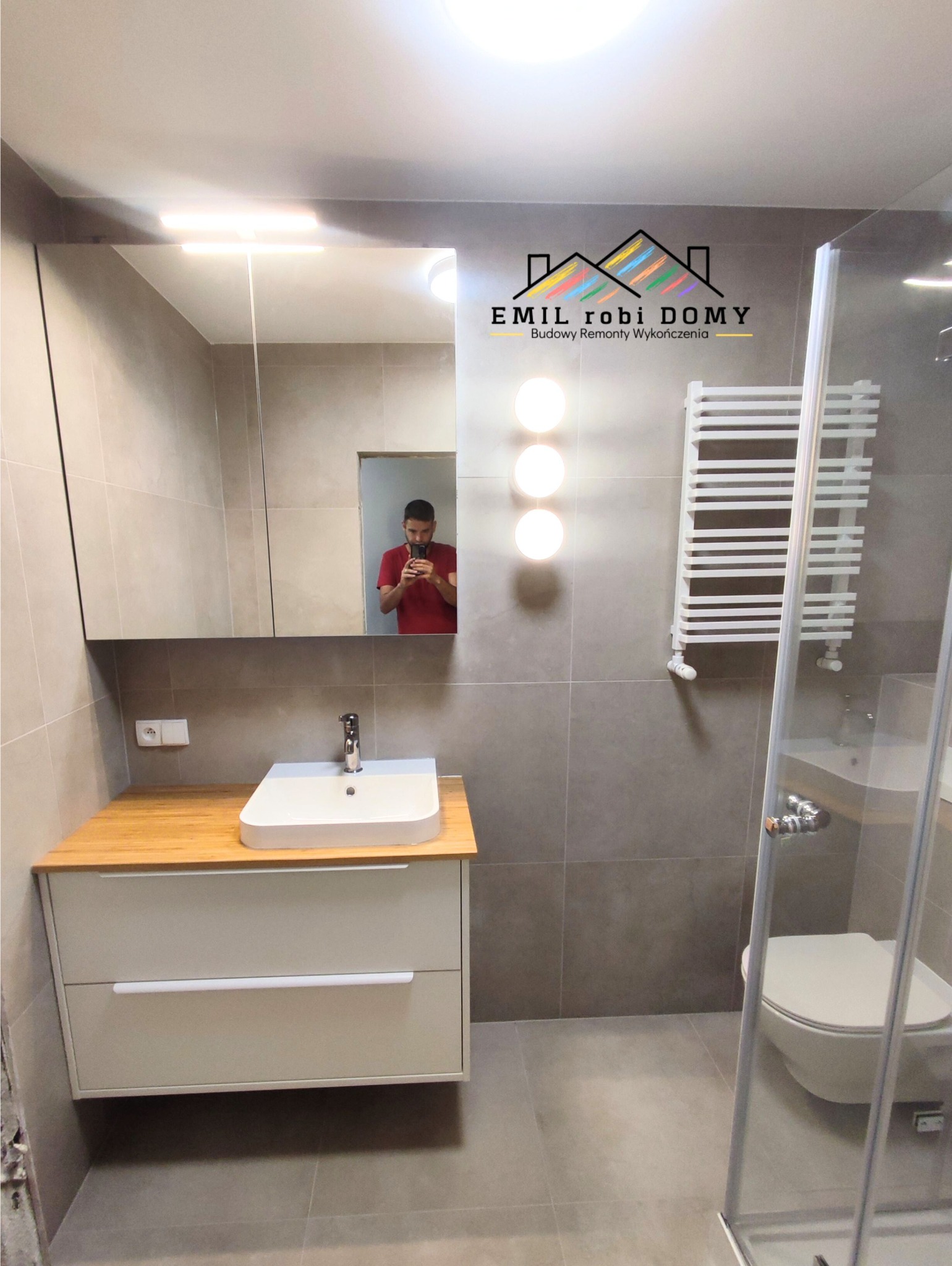 Wykończona łazienka w szarościach z drewnianym blatem pod umywalką, kabiną prysznicową z widoczną toaletą oraz grzejnikiem drabinkowym; w lustrze odbija się osoba robiąca zdjęcie.