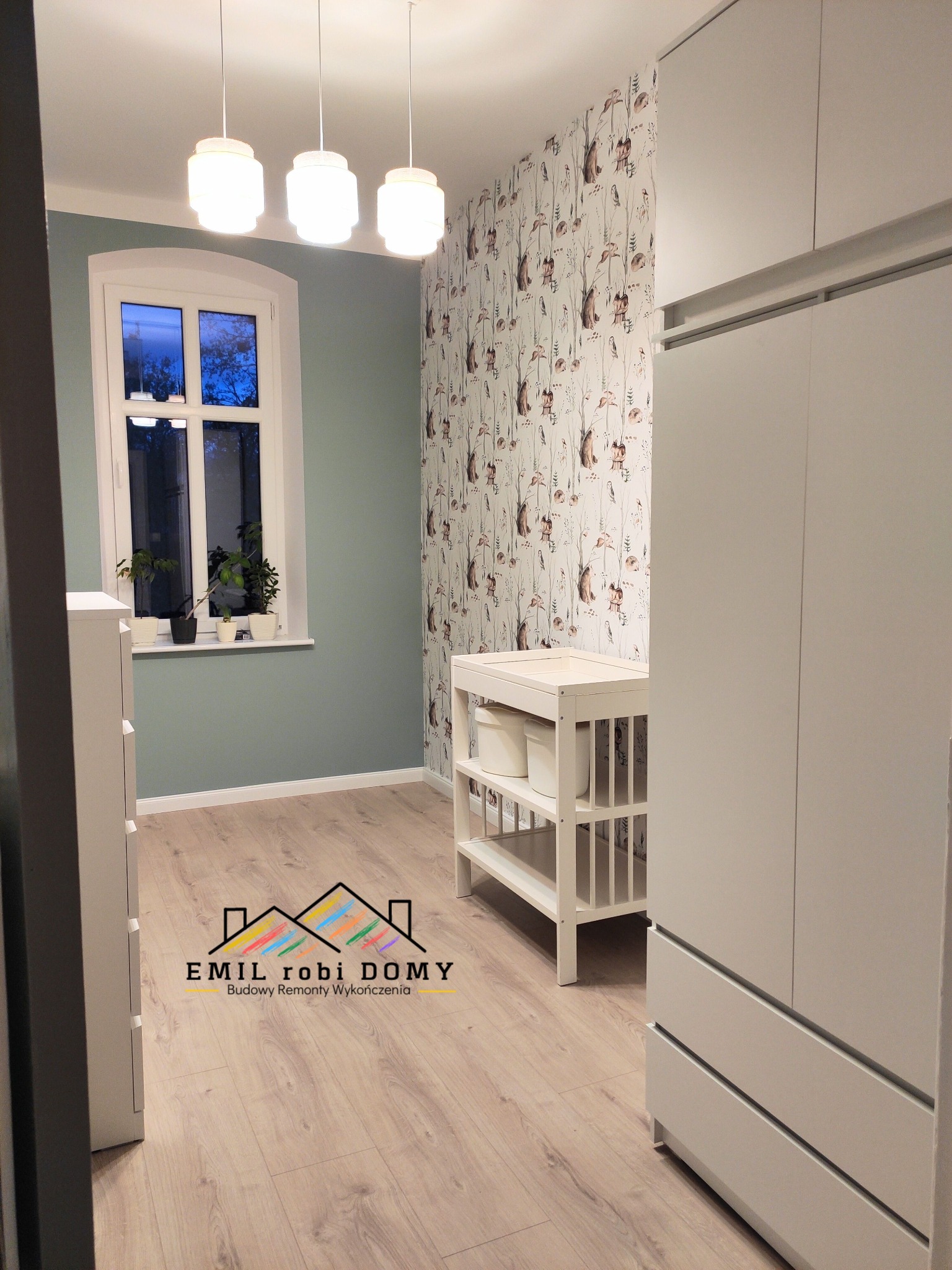 Pokój dziecięcy po remoncie: ściana pomalowana na zielono, tapeta w leśne zwierzęta, białe meble, trzy lampy wiszące nad oknem z roślinami.