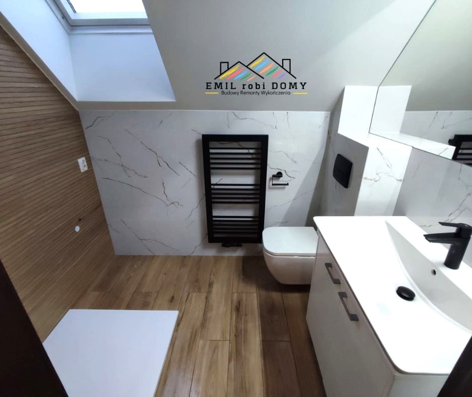 Nowoczesna łazienka na poddaszu ze skośnym sufitem i oknem dachowym, wykończona płytkami imitującymi marmur, drewnopodobną podłogą, z czarnym grzejnikiem drabinkowym i białą szafką umywalkową.
