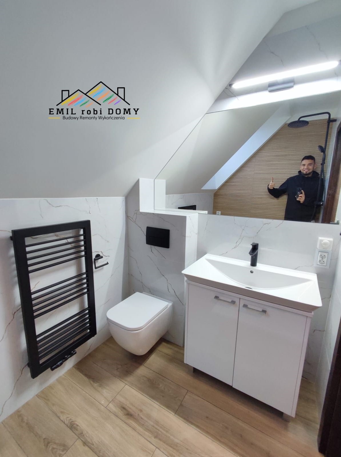 Nowoczesna łazienka na poddaszu z białymi marmurkowymi płytkami, drewnianą podłogą i czarnymi akcentami, w lustrze odbija się uśmiechnięty mężczyzna dający kciuk w górę.