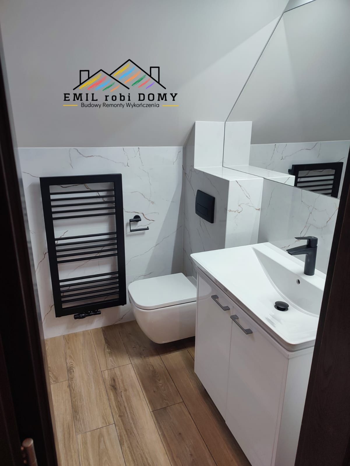 Nowoczesna łazienka w skosach z marmurowymi płytkami, drewnopodobną podłogą, białą zabudową umywalki i czarnym grzejnikiem drabinkowym; widoczne logo firmy 'EMIL robi DOMY'.
