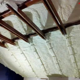 Poddasze ocieplone pianą poliuretanową, widoczne drewniane krokwie i metalowe łączniki, nierównomierna struktura piany.