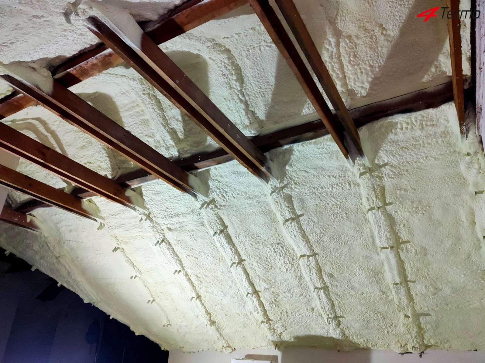 Poddasze ocieplone pianą poliuretanową, widoczne drewniane krokwie i metalowe łączniki, nierównomierna struktura piany.
