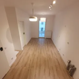 Puste, jasne pomieszczenie z drewnianą podłogą, białymi ścianami i sufitem, widoczne lampy sufitowe, drzwi i grzejnik, przygotowane do dalszych prac wykończeniowych.