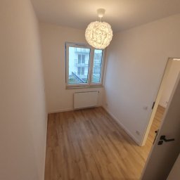 Łukasz Łęszczak - Pusty pokój po remoncie z panelami podłogowymi w odcieniach brązu, świeżo pomalowanymi ścianami na biało i widokiem na balkon przez okno. Nowoczesna lampa wisząca z białym kloszem w kształcie kuli.