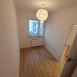 Pusty pokój po remoncie z panelami podłogowymi w odcieniach brązu, świeżo pomalowanymi ścianami na biało i widokiem na balkon przez okno. Nowoczesna lampa wisząca z białym kloszem w kształcie kuli.