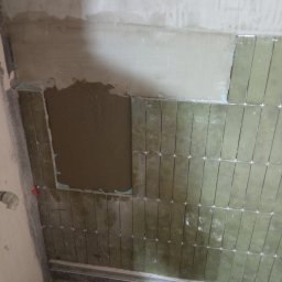 Łukasz Łęszczak - Ściana w trakcie układania podłużnych, oliwkowych płytek z użyciem krzyżyków dystansowych, widoczny fragment ściany z nałożonym klejem oraz elementy montażowe przy podłodze.