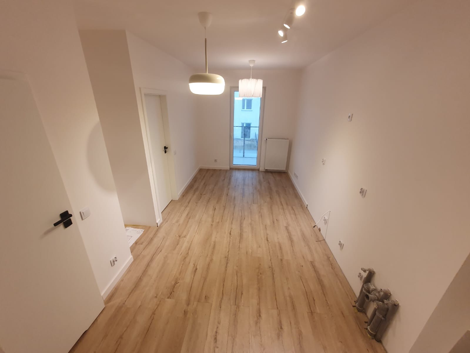 Puste, jasne pomieszczenie z drewnianą podłogą, białymi ścianami i sufitem, widoczne lampy sufitowe, drzwi i grzejnik, przygotowane do dalszych prac wykończeniowych.