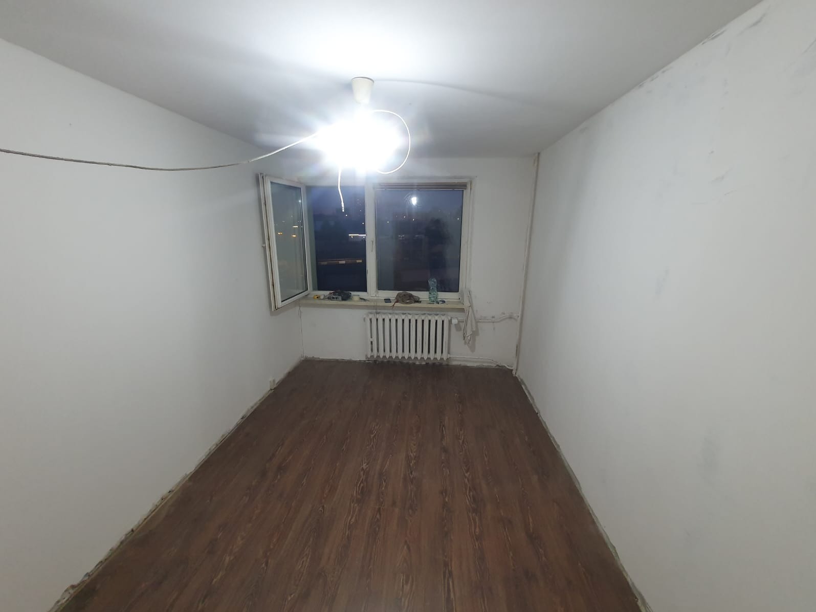 Pusty pokój po remoncie: świeżo pomalowane białe ściany, panele podłogowe w odcieniach brązu, grzejnik pod oknem, surowa żarówka zwisająca z sufitu.