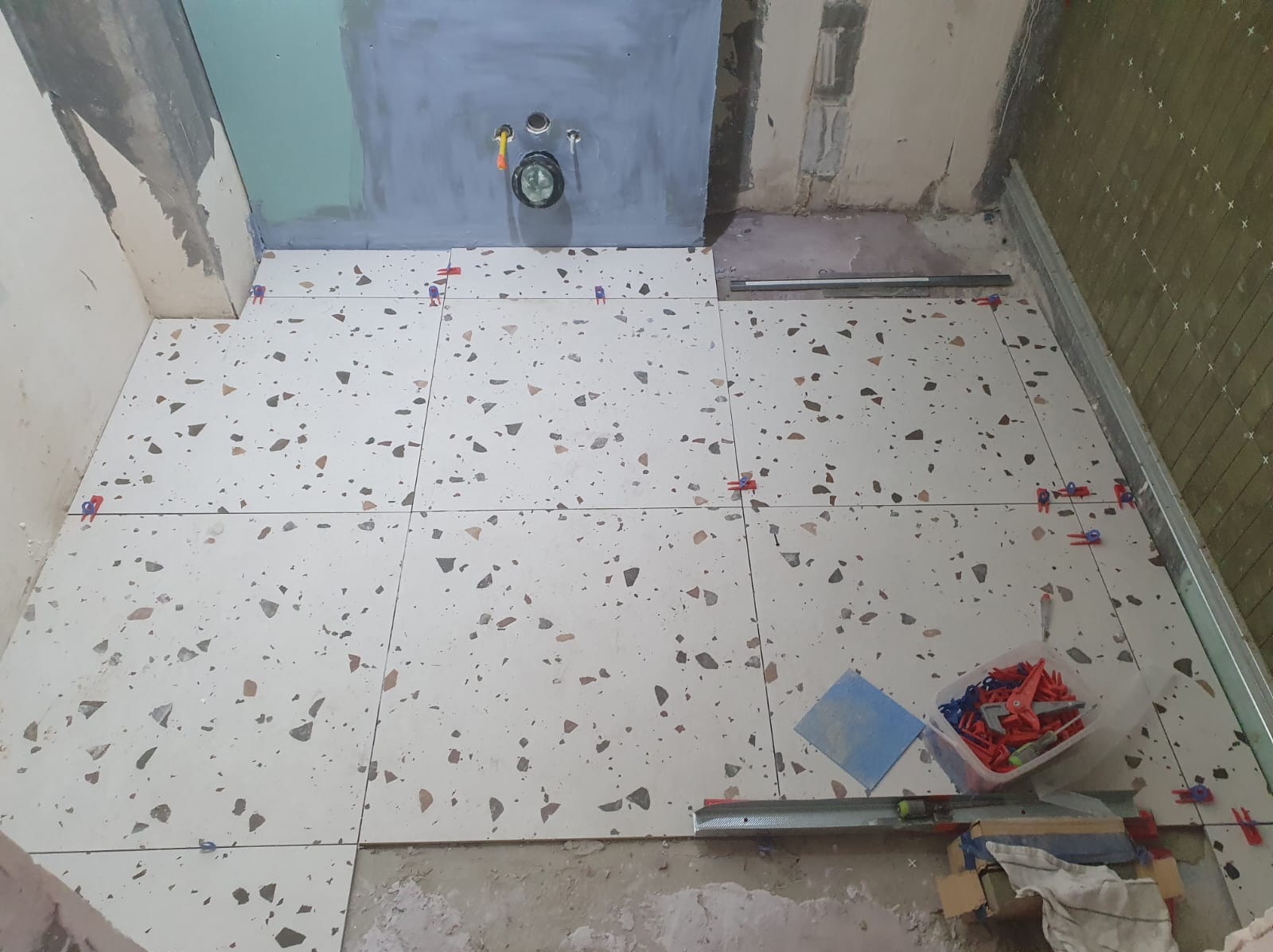 Układanie płytek podłogowych z wzorem lastryko w łazience, widoczne klipsy poziomujące i narzędzia glazurnicze, ściany przygotowane pod wykończenie.