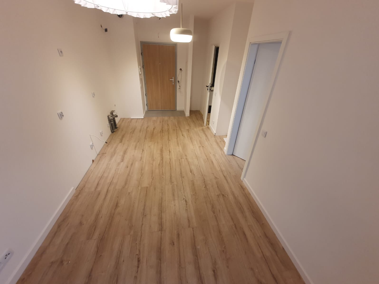 Korytarz po remoncie z nową podłogą z paneli imitujących drewno, widoczne niedokończone prace elektryczne na ścianie po lewej stronie, białe ściany i drzwi, sufit z lampą.