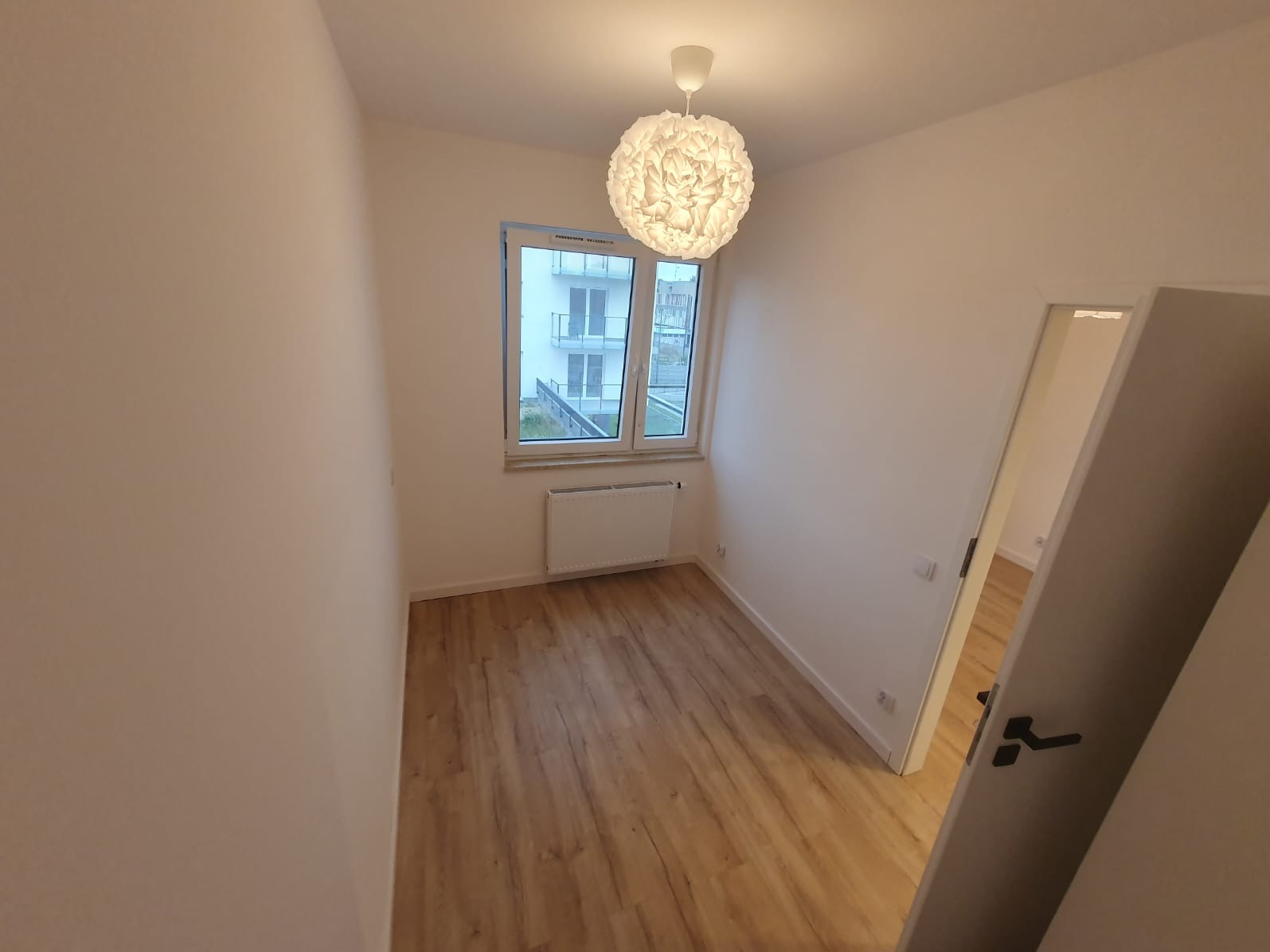 Pusty pokój po remoncie z panelami podłogowymi w odcieniach brązu, świeżo pomalowanymi ścianami na biało i widokiem na balkon przez okno. Nowoczesna lampa wisząca z białym kloszem w kształcie kuli.