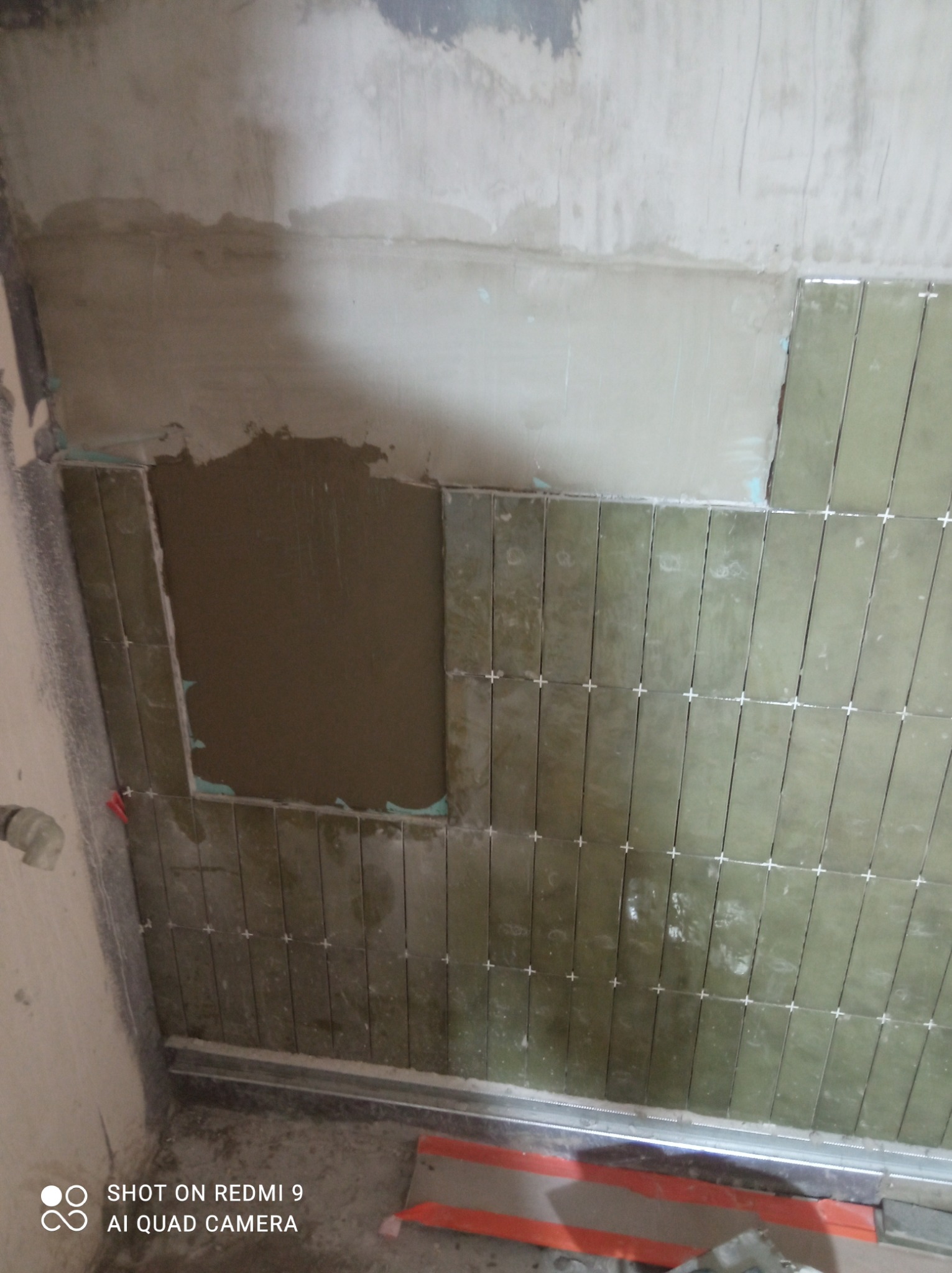 Ściana w trakcie układania podłużnych, oliwkowych płytek z użyciem krzyżyków dystansowych, widoczny fragment ściany z nałożonym klejem oraz elementy montażowe przy podłodze.