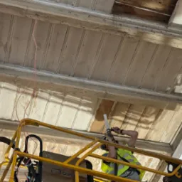 Pracownik na podnośniku w kamizelce odblaskowej, za pomocą narzędzia usuwa starą farbę z drewnianego sufitu z dekoracyjnymi listwami, widoczne ślady wcześniejszych prac i odsłonięte fragmenty...