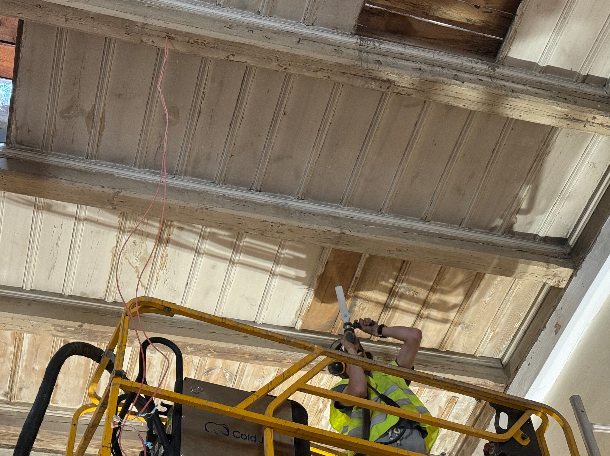 Pracownik na podnośniku w kamizelce odblaskowej, za pomocą narzędzia usuwa starą farbę z drewnianego sufitu z dekoracyjnymi listwami, widoczne ślady wcześniejszych prac i odsłonięte fragmenty...