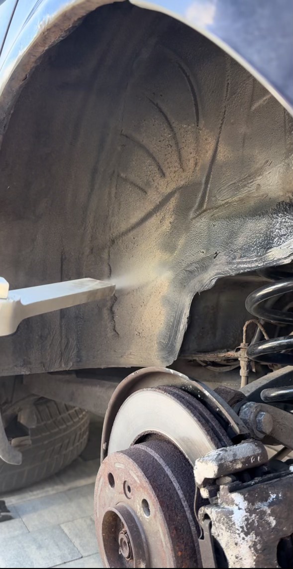 Piaskowanie podwozia samochodu w trakcie pracy, widoczny strumień piasku i fragment tarczy hamulcowej z rdzawym nalotem.