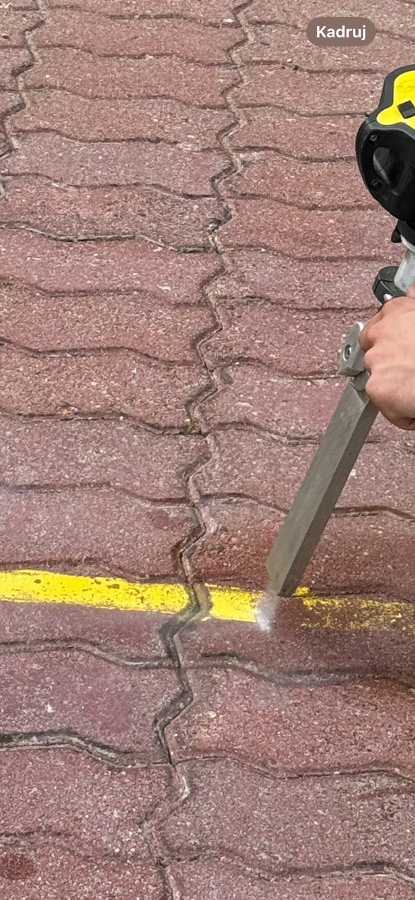 Czerwona kostka brukowa z żółtą linią poddawana obróbce strumieniowo-ściernej, widoczny pył i fragment urządzenia.