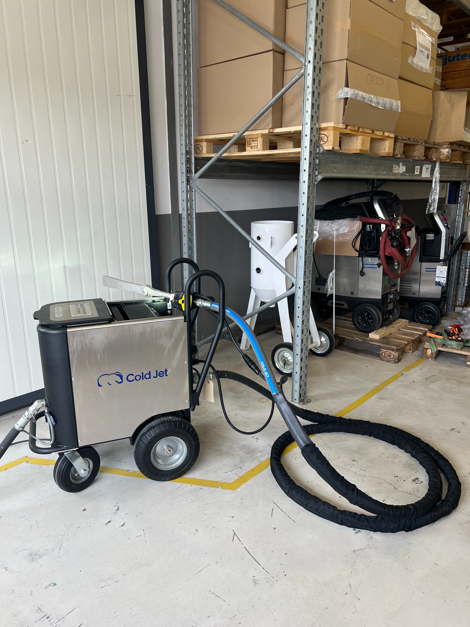 Mobilna maszyna do czyszczenia suchym lodem Cold Jet z niebieskim wężem, stojąca na betonowej podłodze magazynu z żółtymi liniami, w tle metalowe regały z kartonami.
