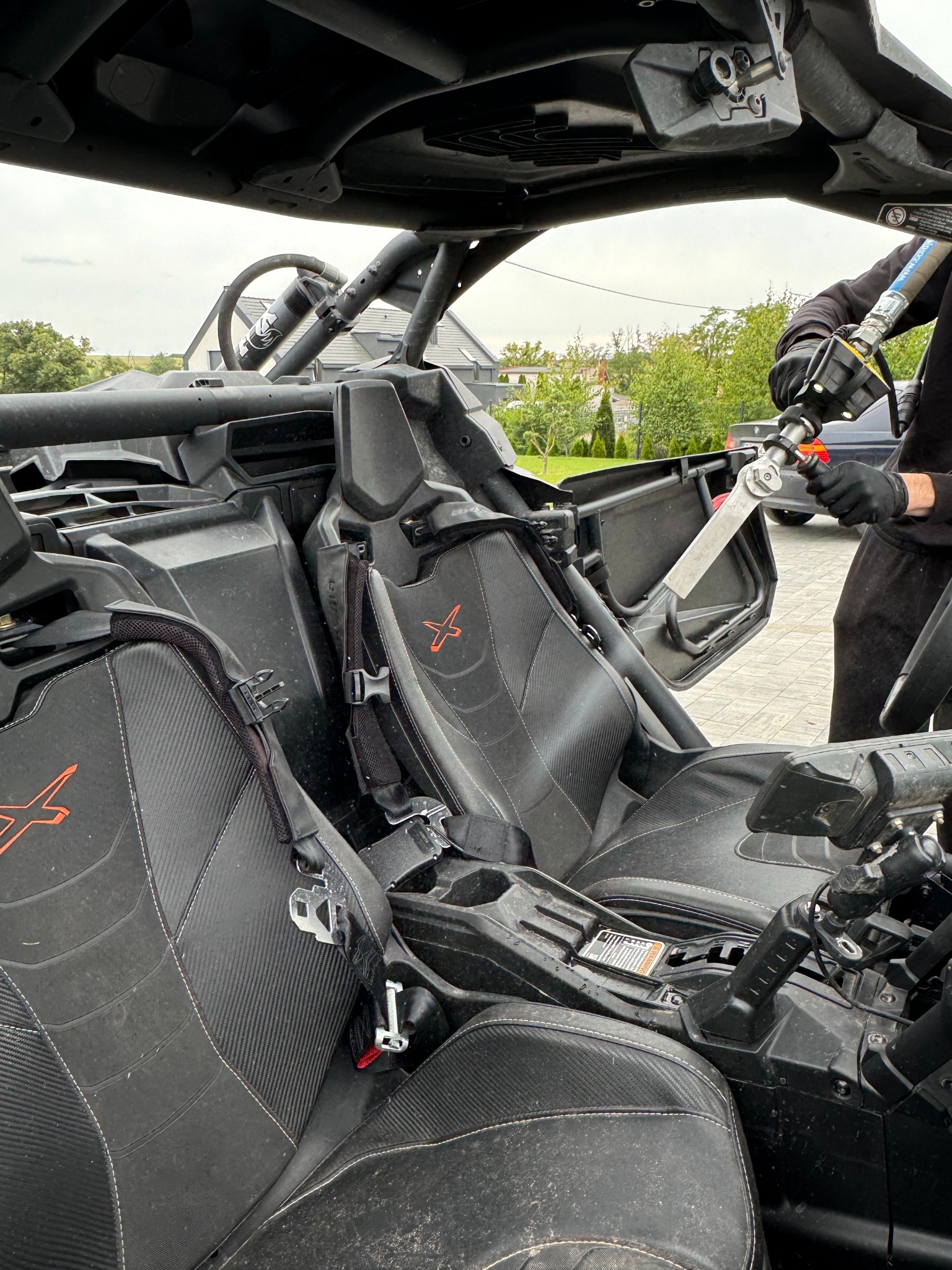 Wnętrze brudnego, czarnego pojazdu terenowego typu buggy z widocznymi siedzeniami, pasami bezpieczeństwa i elementami konstrukcyjnymi, czyszczone lancą ciśnieniową przez osobę w czarnych rękawicach.