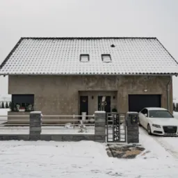 Surowy, nowoczesny dom z płaskim dachem pokrytym śniegiem, z szarym ogrodzeniem i białym samochodem na ośnieżonym podjeździe w zimowy dzień.