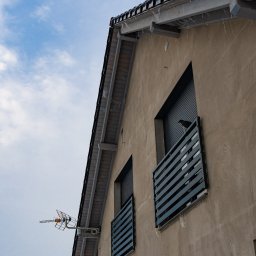 EKO - DOM - Fragment elewacji domu z balkonami z poziomymi barierkami, na jednym z balkonów siedzi ptak, na dachu widoczna antena telewizyjna.